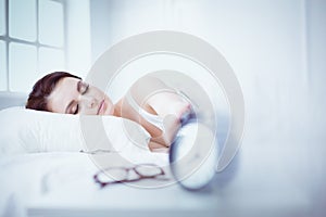Beautiful young woman sleeping while lying in bed comfortably and blissfully on the background of alarm clock