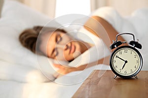 Beautiful young woman sleeping and smiling while lying in bed comfortably and blissfully on the background of alarm