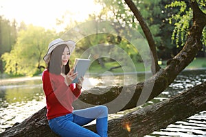 Beautiful young woman with smile reading book in the park and sitting on a wooden tree on river