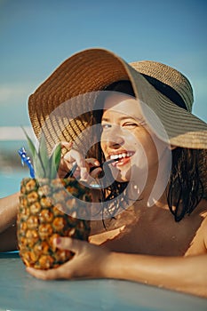 Beautiful young woman smiling and drinking cocktail in pineapple, relaxing in pool on summer vacation. Girl in hat enjoying warm