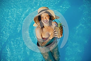 Beautiful young woman smiling, holding cocktail in pineapple and relaxing in pool, summer vacation. Girl in hat enjoying warm