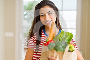 Beautiful young woman smiling holding a paper bag full of fresh groceries at home