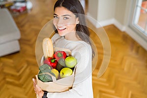 Beautiful young woman smiling holding a paper bag full of fresh groceries at home