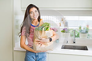 Beautiful young woman smiling holding a paper bag full of groceries at the kitchen