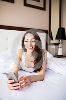 Beautiful young woman smiling and holding smart phone while lying in the bed at home