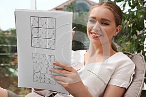 Beautiful young woman solving sudoku puzzle near window indoors, focus on hand