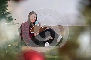 A beautiful young woman spends her Christmas holiday relaxing, enjoying tea and reading a book on the sofa in the living
