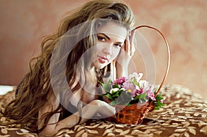 Beautiful young woman with spring flowers and long wavy hair lying on bedroom, beauty portrait, interior