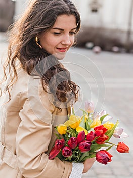 Beautiful young woman with spring tulips flowers bouquet at city street. Happy girl walking outdoors. Spring portrait of
