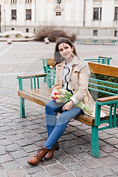 Beautiful young woman with spring tulips flowers bouquet at city street. Happy girl sitting on a bench outdoors. Spring portrait