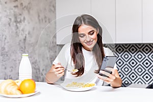 Beautiful young woman standing by kitchen counter with bowl of fresh berries and reading text message on her cell phone. Female