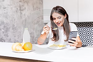 Beautiful young woman standing by kitchen counter with bowl of fresh berries and reading text message on her cell phone. Female