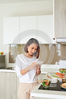 Beautiful young woman standing by kitchen counter reading text message on her cell phone