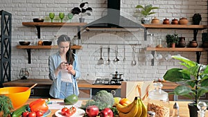 A beautiful young woman is standing in the kitchen and texting with her beloved husband using her smart phone.