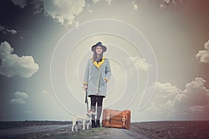 beautiful young woman standing near dog and suitcase on the wonderful road on the field