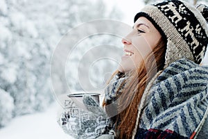 Beautiful young woman standing among snowy trees in winter forest and enjoying cup of hot tea