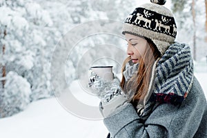 Beautiful young woman standing among snowy trees in winter forest and enjoying cup of hot tea