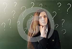 Beautiful young woman stands in thought on background of chalk board and question marks. Education concept