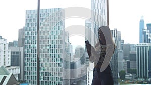 Beautiful young woman stands by the window in aparment uses smart phone on background of urban city. 3840x2160