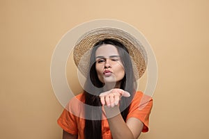 Beautiful young woman in straw hat blowing kiss on beige background