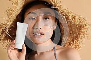 Beautiful young woman in straw hat with sunscreen on her face holding sun protection cream against beige background, closeup