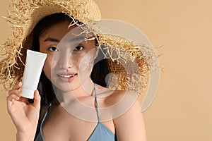 Beautiful young woman in straw hat with sunscreen on her face holding sun protection cream against beige background, space for