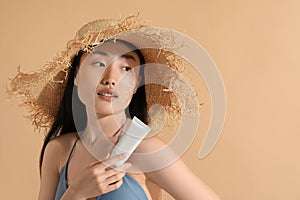 Beautiful young woman in straw hat with sunscreen on her face holding sun protection cream against beige background, space for