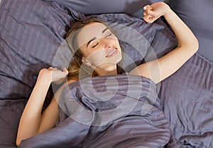 Beautiful young woman stretching after waking up while in cozy bedroom bed, smiling rested girl feeling refreshed sitting on white