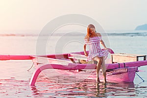 A beautiful young woman in a striped sundress wetting her feet in the water, sitting on a boat in the open ocean. View from the