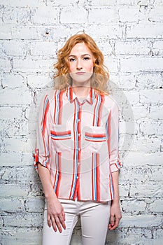 A beautiful young woman student with red curly hair and freckles is leaning against a brick wall of gray color. Dressed in a red s