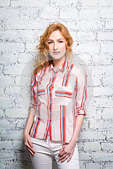 A beautiful young woman student with red curly hair and freckles is leaning against a brick wall of gray color. Dressed in a red s