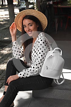 Beautiful young woman with stylish backpack and hat sitting on stairs outdoors