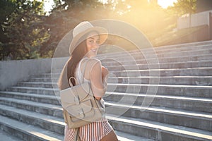 Beautiful young woman with stylish backpack and hat on stairs outdoors