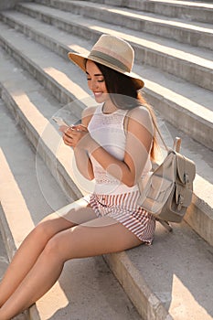 Beautiful young woman with stylish backpack and smartphone sitting on stairs outdoors