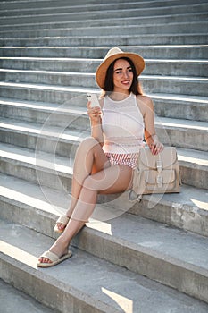 Beautiful young woman with stylish backpack and smartphone sitting on stairs outdoors