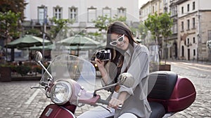 Beautiful young woman in stylish glasses holding vintage camera and sitting on the modern scooter in the middle of the