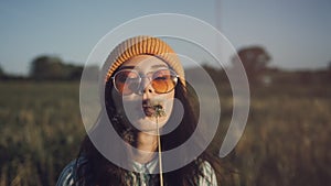 A beautiful young woman in stylish sunglasses stand in a field and blow a dandelion. A smiling hipster girl wearing