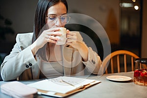 Beautiful young woman in suit writing her business plans while drinking coffee on break time