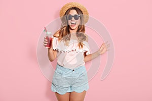 Beautiful young woman in summer blouse, short, hat and sunglasses, holding jug with cold beverage while standing on pink