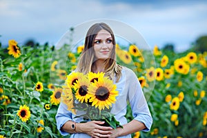 Beautiful young woman on sunflower field with bouquet flowers. Happy girl on summer sunset day.