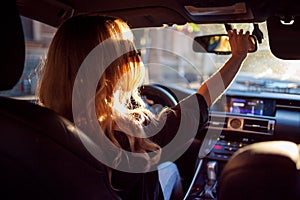 Beautiful young woman in sunglasses sitting behind the wheel of a car and corrects mirror. Photo from the back seat