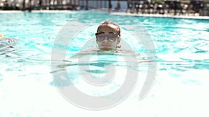 Beautiful young woman in sunglasses swimming and comes up on pool edge by hands and joyfully looking at camera.