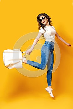 Beautiful young woman in sunglasses, white shirt, blue jeans jumping with bag on the yellow background