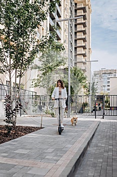 Beautiful young woman in sunglasses and white suit is riding on her electric scooter with Corgi dog near modern building in city