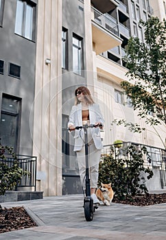 Beautiful young woman in sunglasses and white suit is riding on her electric scooter with Corgi dog near modern building in city