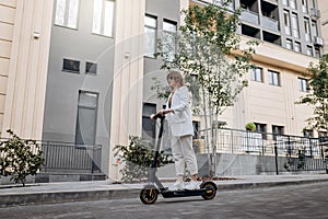 Beautiful young woman in sunglasses and white suit is riding on her electric scooter near modern building in city and looking away