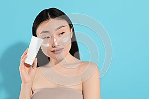 Beautiful young woman with sunscreen on her face holding sun protection cream against light blue background, space for text