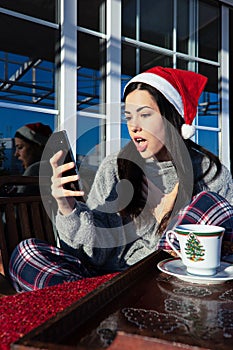 Beautiful young woman surprised while using her mobile phone wearing a Santa Claus hat on Christmas