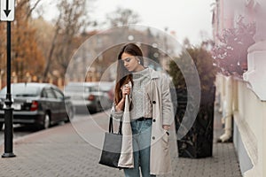 Beautiful young woman in a sweater in a fashionable long light trench coat in jeans with a black leather bag standing on a street