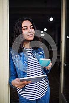 A beautiful young woman with tablet and coffee standing at the front door in cafe.